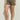 Wearing Sanctuary Clothing's fable short in vintage olive, paired with a black top, studded brown belt, and black strappy sandals, a person stands against a light wall. Only the lower half of their body is visible.