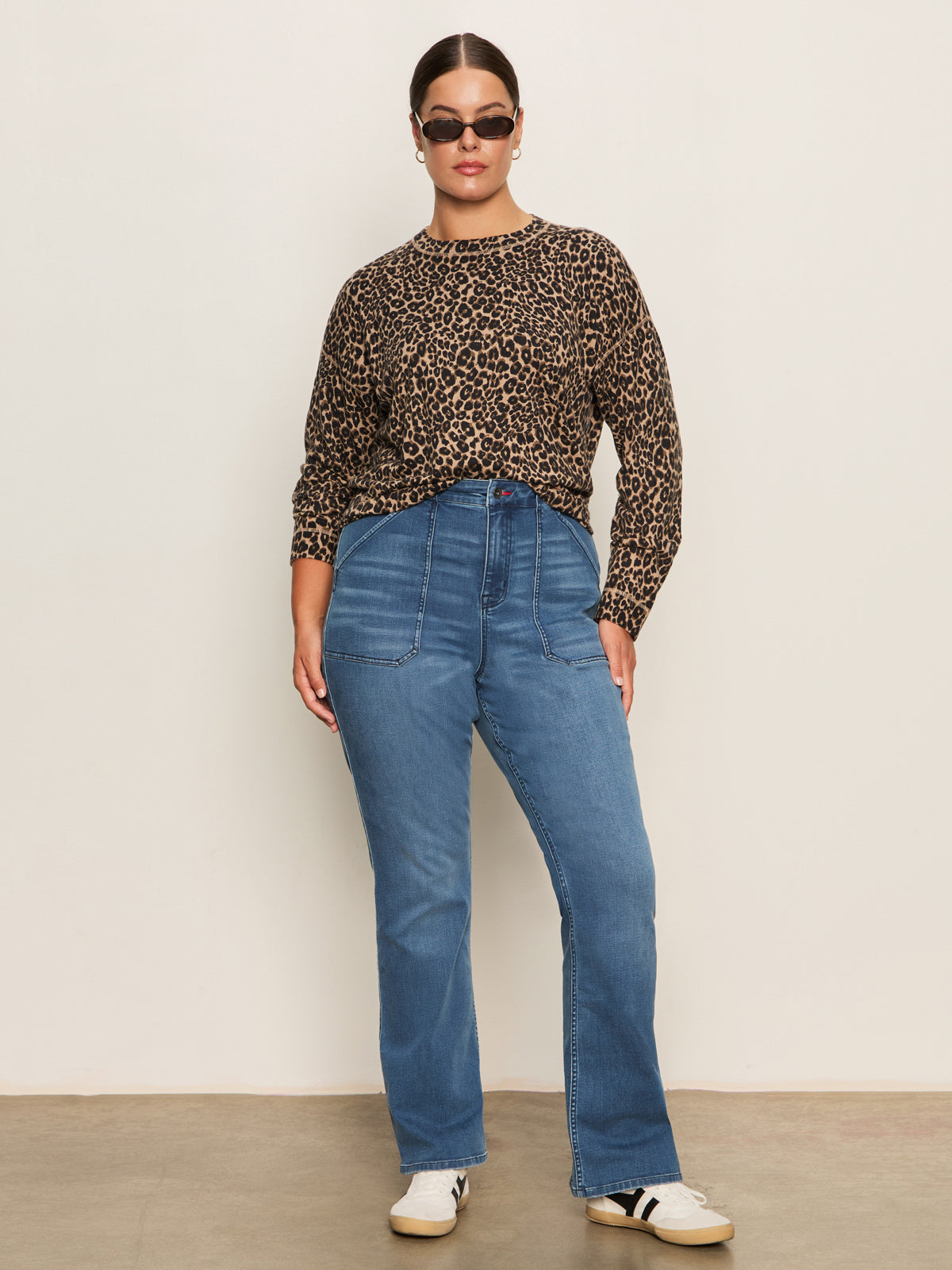 A woman stands against a plain wall wearing sunglasses, a long-sleeve leopard print top, and Sanctuary Clothing sculpted hayden bootcut jeans in lucky streak (extended sizing), paired with white sneakers with black stripes.