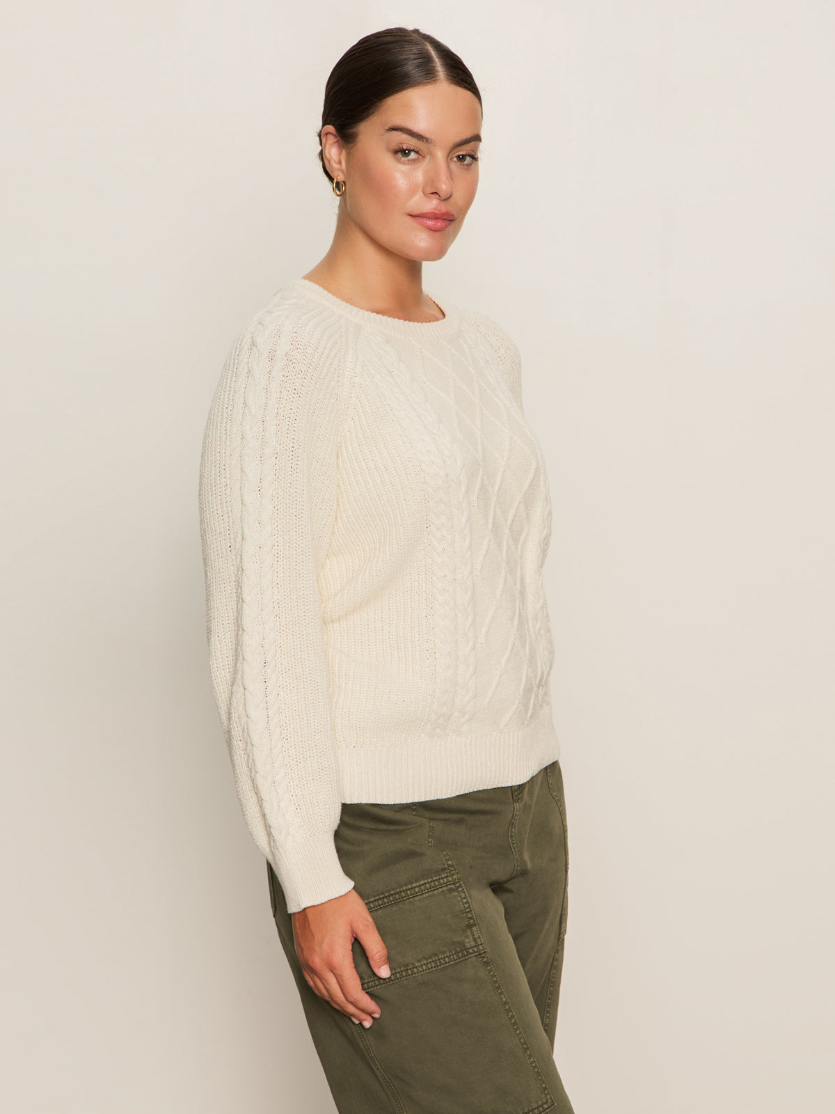 A woman with dark hair tied back is wearing the Sanctuary Clothing nomad cable popover chalk (extended sizing), standing against a light beige background and looking at the camera.