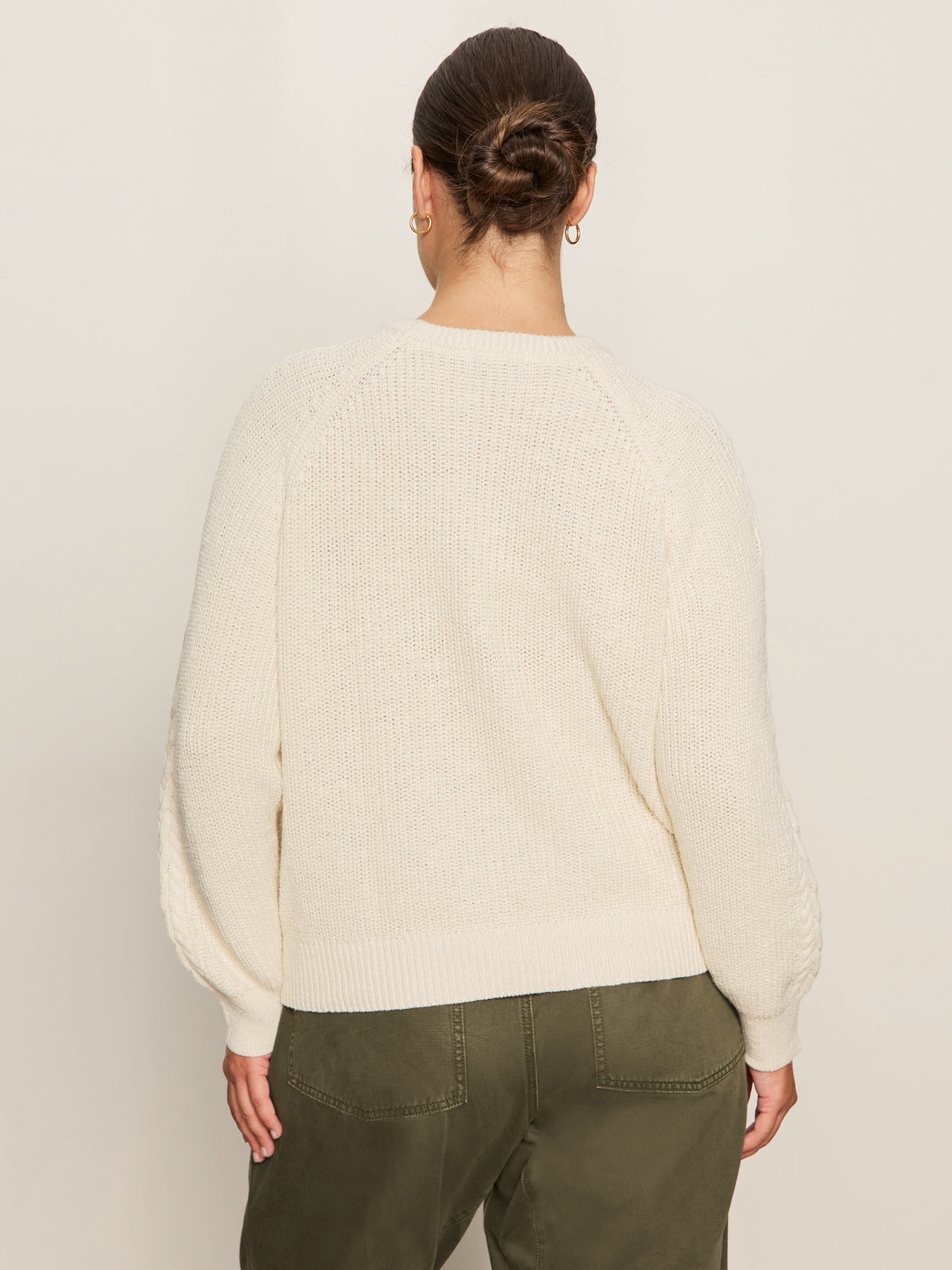 A person with dark hair in a bun is shown from the back, wearing Sanctuary Clothing's nomad cable popover chalk extended sizing sweater, featuring textured sleeves, paired with olive green pants against a neutral background.