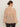 A woman with dark hair in a low bun is seen from behind wearing Sanctuary Clothingžs fade into you ruffle blouse in fawn�a long-sleeve, loose, sheer beige top with a wide back ruffle�paired with dark pants.