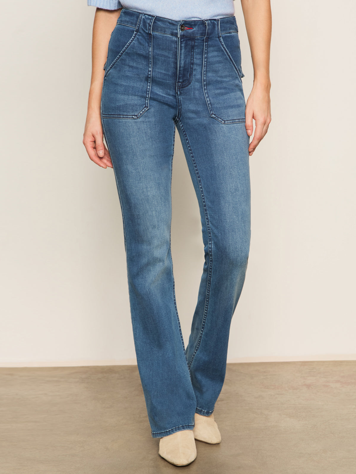 A person wears Sanctuary Clothing's sculpted hayden bootcut lucky streak jeans, featuring high-waisted blue denim with front patch pockets and white stitching, standing against a plain light backdrop, hands relaxed at their sides.