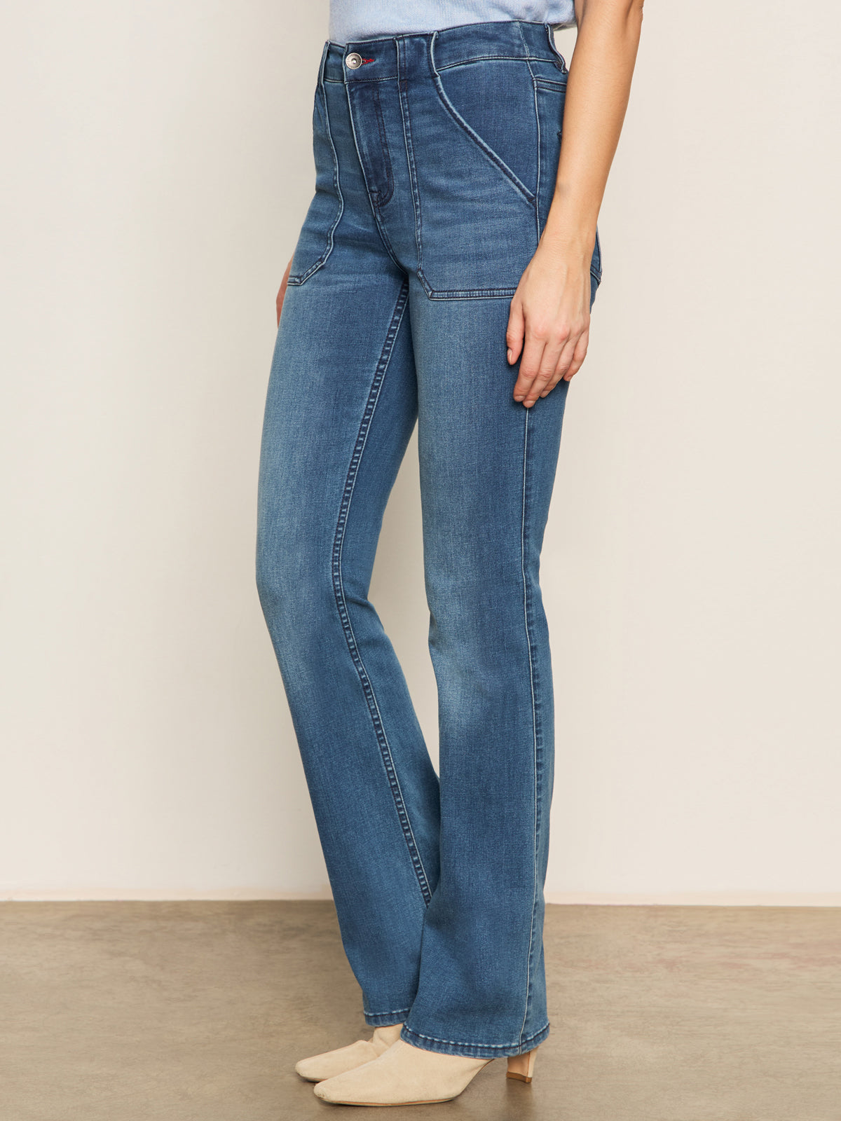 A person stands indoors in Sanctuary Clothing's sculpted hayden bootcut lucky streak jeans and beige pointed-toe shoes; only their upper body is partially visible against a neutral wall background.