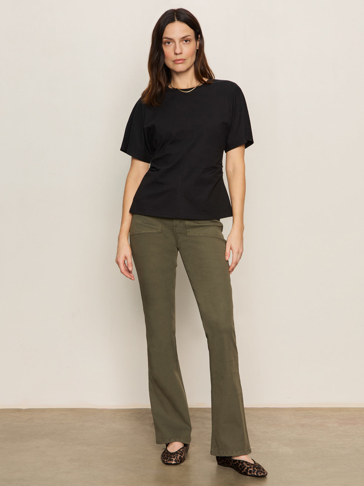 A woman with long brown hair and a neutral expression stands against a plain light-colored wall, wearing Sanctuary Clothing's sculpted brook bootcut pant in kalamata, a black short-sleeve top, and leopard print flats.