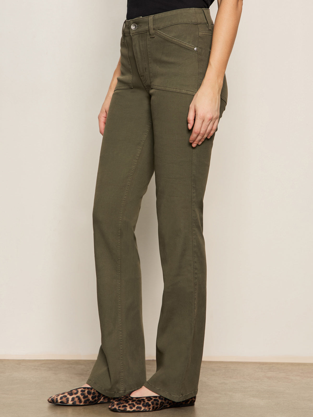 A person stands against a plain light wall, arms relaxed by their side, wearing Sanctuary Clothing's sculpted brook bootcut pant in kalamata and leopard print flats.