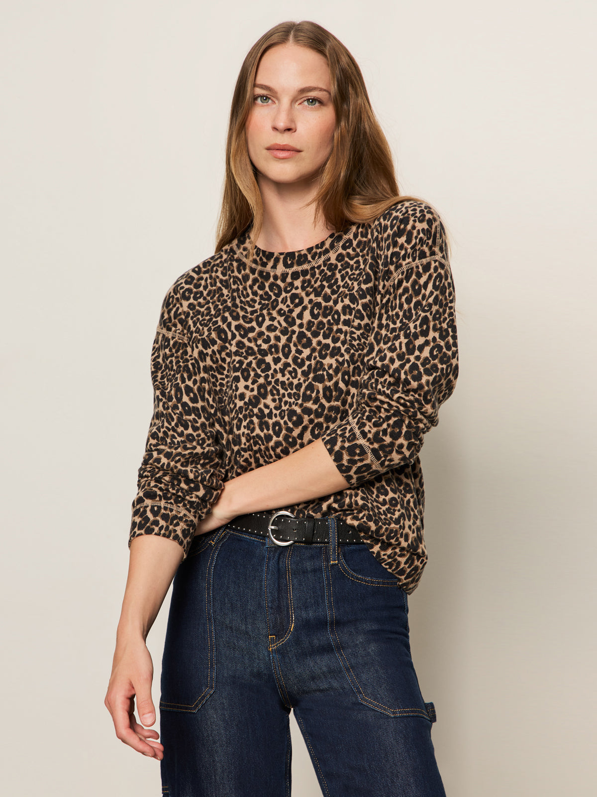 A woman with long brown hair wears the Sanctuary Clothing 24 7 popover french kitty sweater, tucked into high-waisted dark blue jeans, posing against a plain light background.