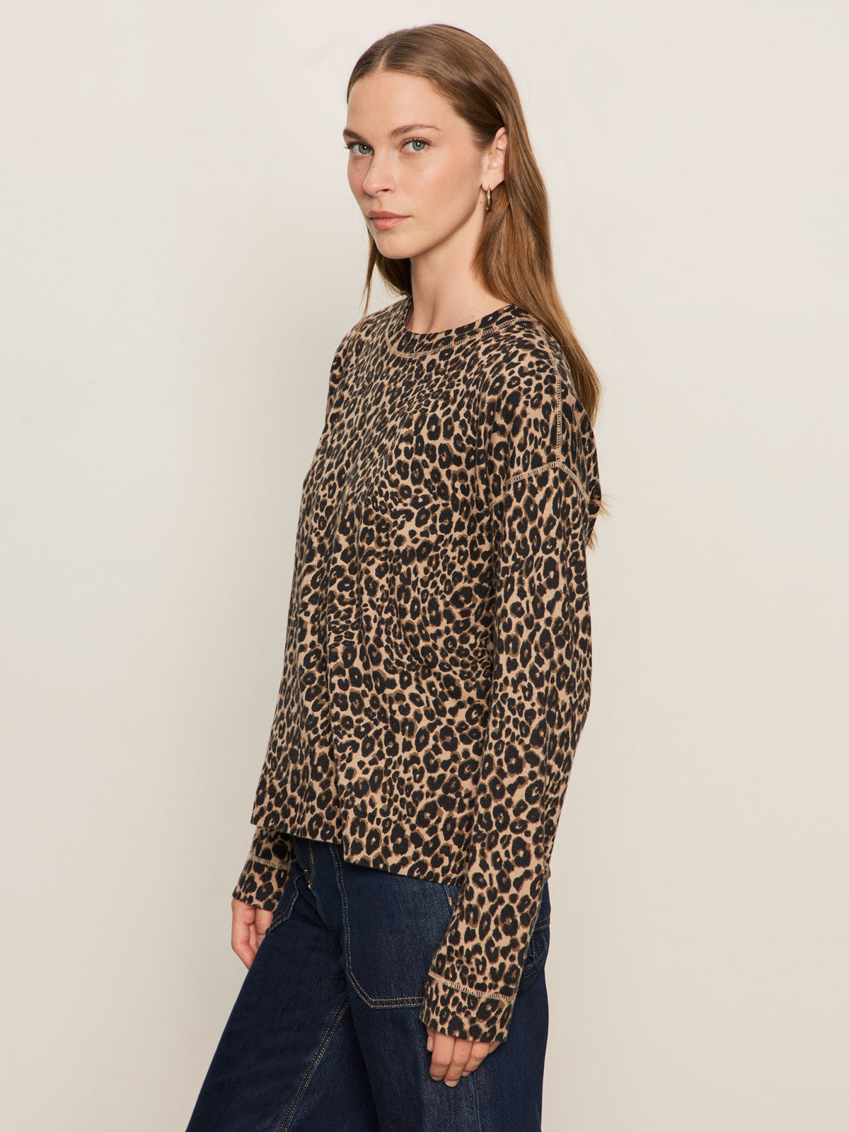 A woman with long brown hair models the Sanctuary Clothing "24 7 popover french kitty" leopard print long-sleeve top and dark blue jeans against a light background, looking slightly to the side with a neutral expression.