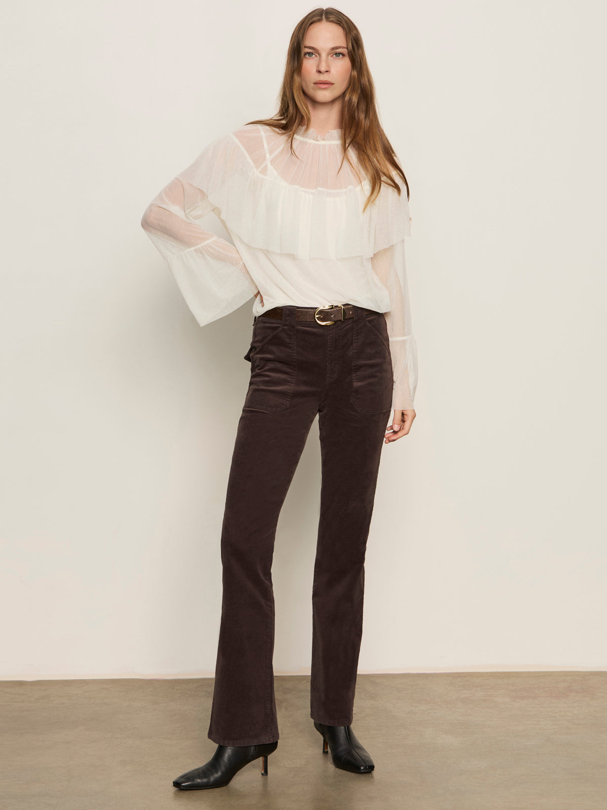 A woman stands against a plain wall wearing the ethereal blouse chalk by Sanctuary Clothing�a sheer white ruffled top�paired with high-waisted dark brown corduroy pants, black heeled boots, and a belt. Her long hair is down, with one hand at her side.