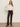 A woman stands against a plain wall wearing the ethereal blouse chalk by Sanctuary Clothing�a sheer white ruffled top�paired with high-waisted dark brown corduroy pants, black heeled boots, and a belt. Her long hair is down, with one hand at her side.