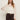A woman with long brown hair wears Sanctuary Clothingžs œethereal blouse chalk��a sheer white ruffled top with flared sleeves�paired with dark high-waisted pants and a belt, standing against a plain light background.