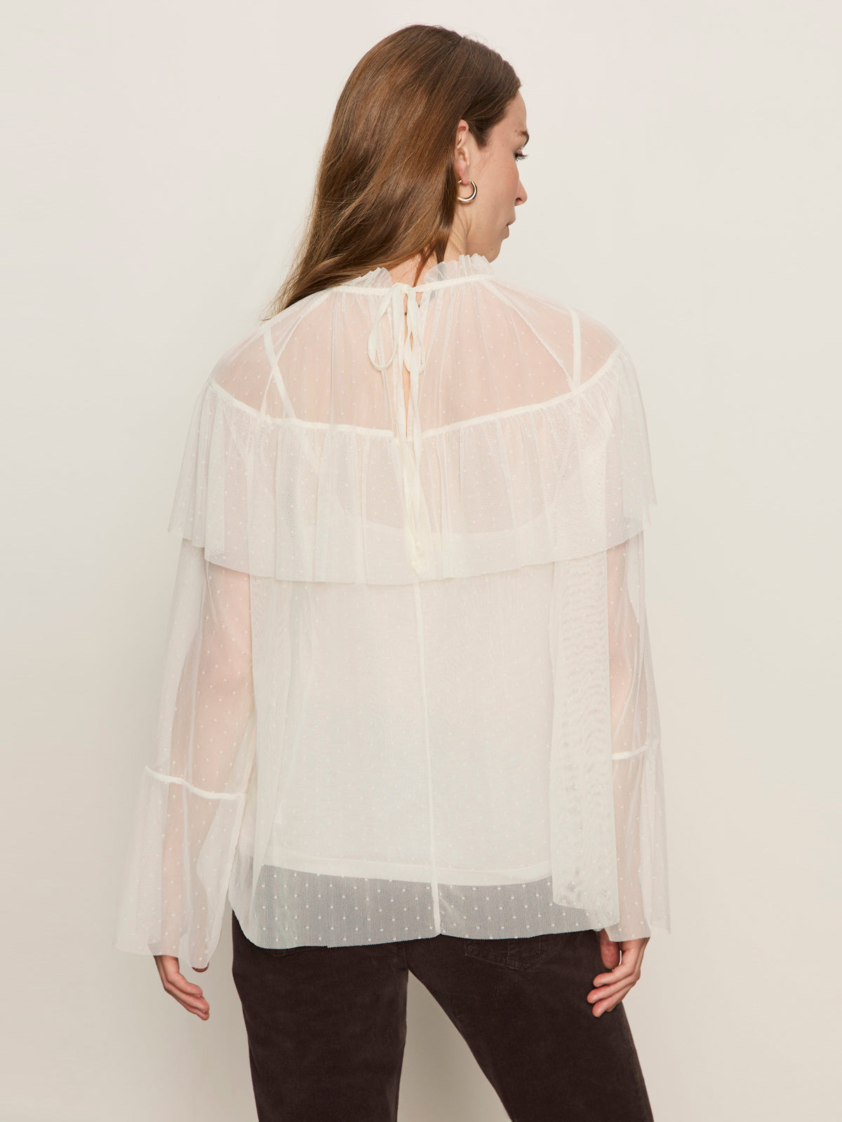 A woman with long brown hair is seen from behind wearing the Sanctuary Clothing "the ethereal blouse chalk," a loose, semi-sheer white top with ruffle details and neck tie, paired with dark pants.
