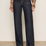 A person wears Sanctuary Clothingžs ekara slim wide jean in indigo dark rinse wash, featuring prominent seams and front buttons, paired with an animal print long-sleeve blouse and black shoes against a plain background.