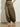 A person wears Sanctuary Clothing's float on balloon pant timber—loose, olive green drawstring pants with elastic cuffs—paired with tan strappy flat sandals, standing on a light brown floor against a plain white wall.