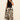 A woman stands against a light wall, wearing the Sanctuary Clothing sandbar mix media midi dress in lotus with black (extended sizing). The dress features bold beige, black, and brown floral patterns. She has curly hair, sandals, and one hand in her pocket.