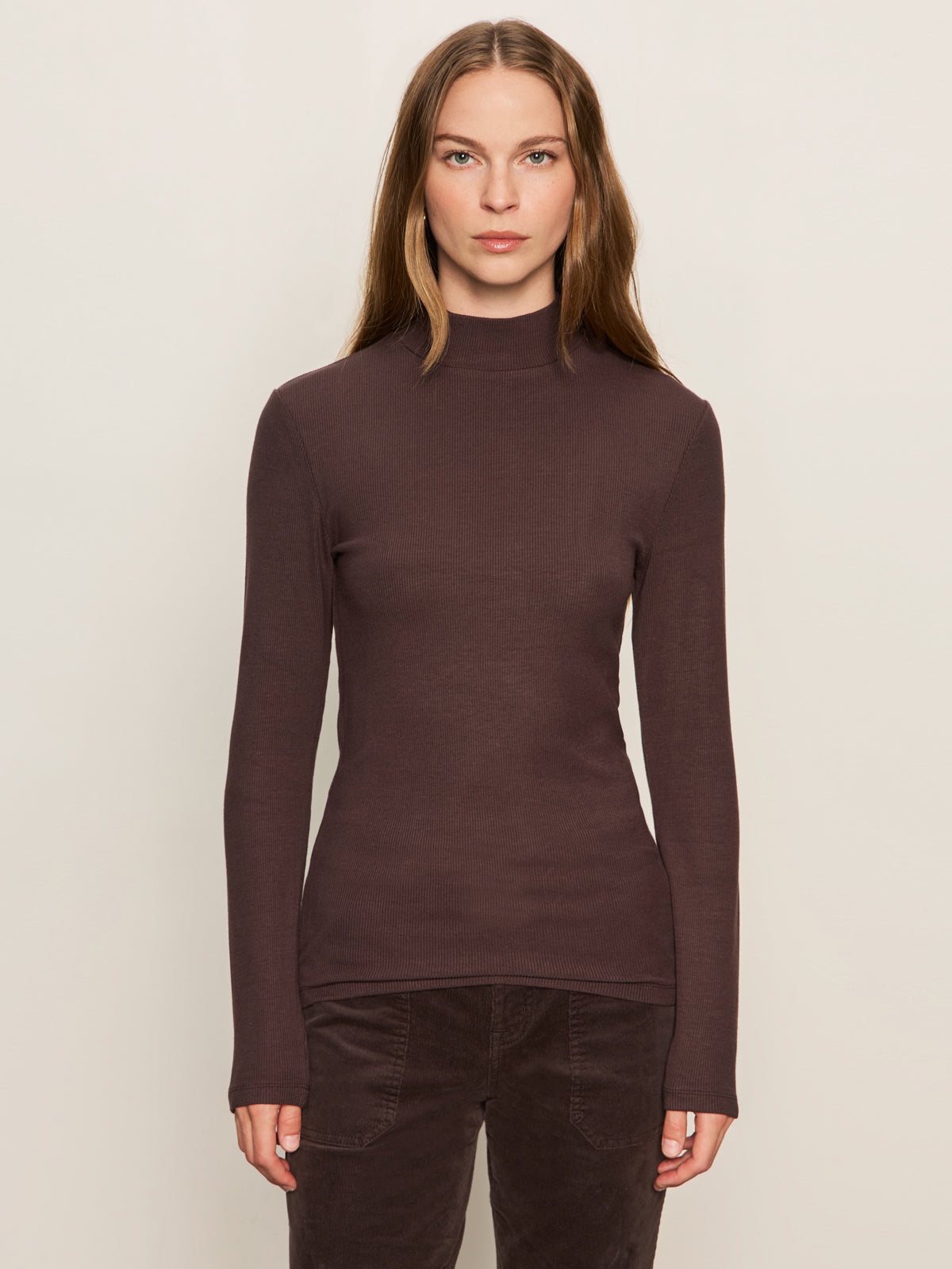 A woman with straight brown hair wears the Sanctuary Clothing ls mock essential top in semi sweet, a fitted long-sleeve high-neck dark brown top, paired with dark pants, standing against a plain light background and facing forward.