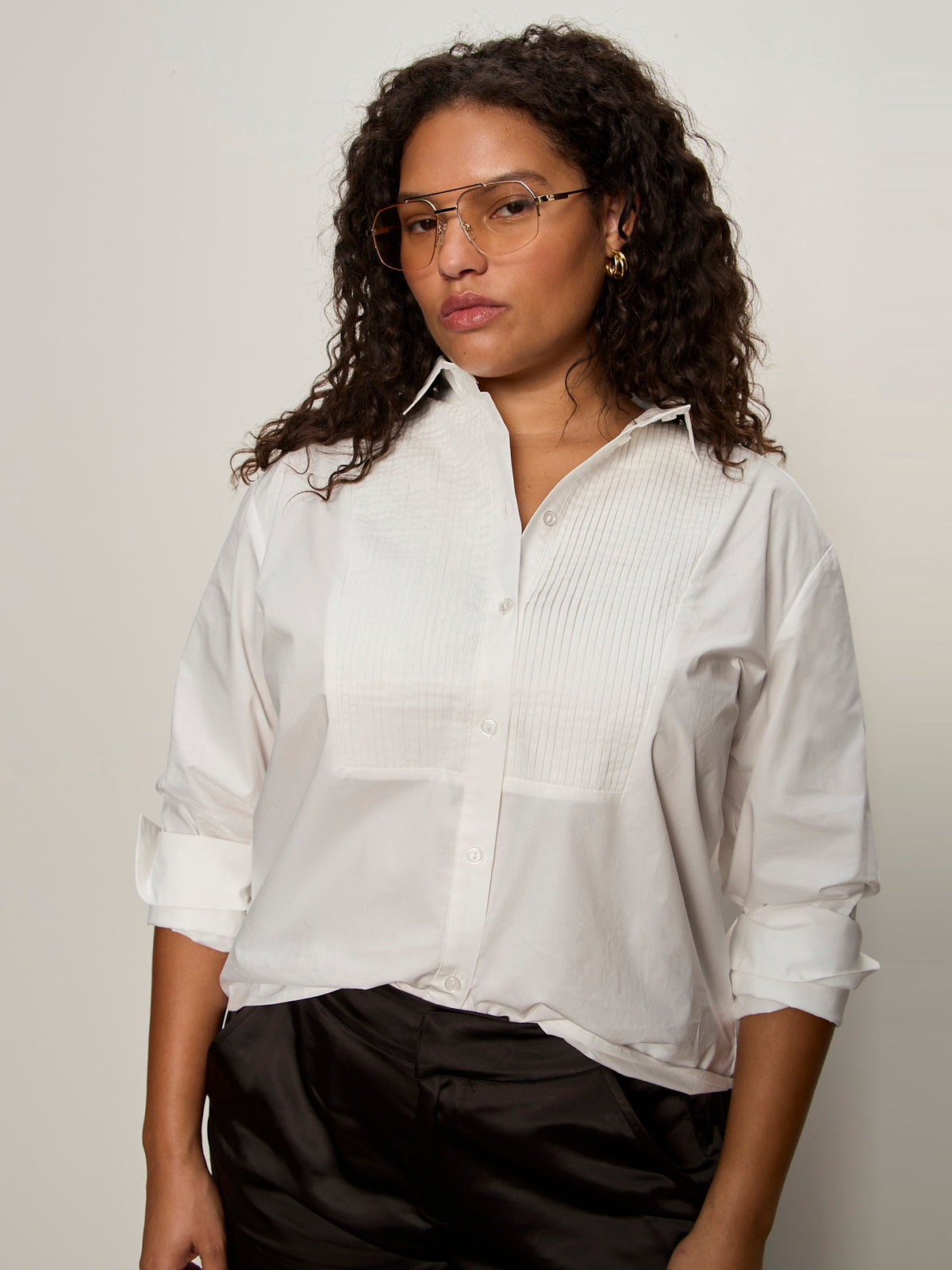 A woman with curly hair, wearing gold-rimmed glasses and black pants, models the Sanctuary Clothing new era tuxedo shirt white extended sizing against a plain light background with a neutral expression.