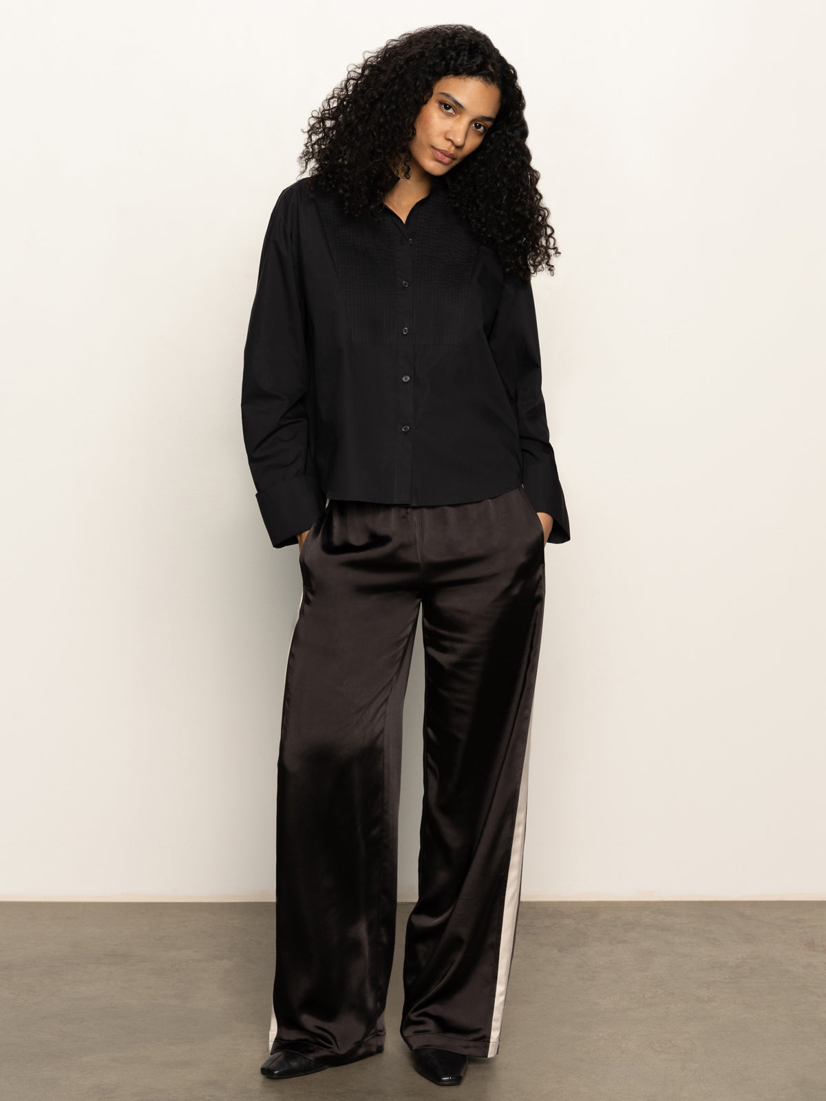 A woman with curly hair stands against a plain wall wearing the Sanctuary Clothing new era tuxedo shirt in black, paired with loose dark brown satin pants featuring a white side stripe. She looks neutral and relaxed.