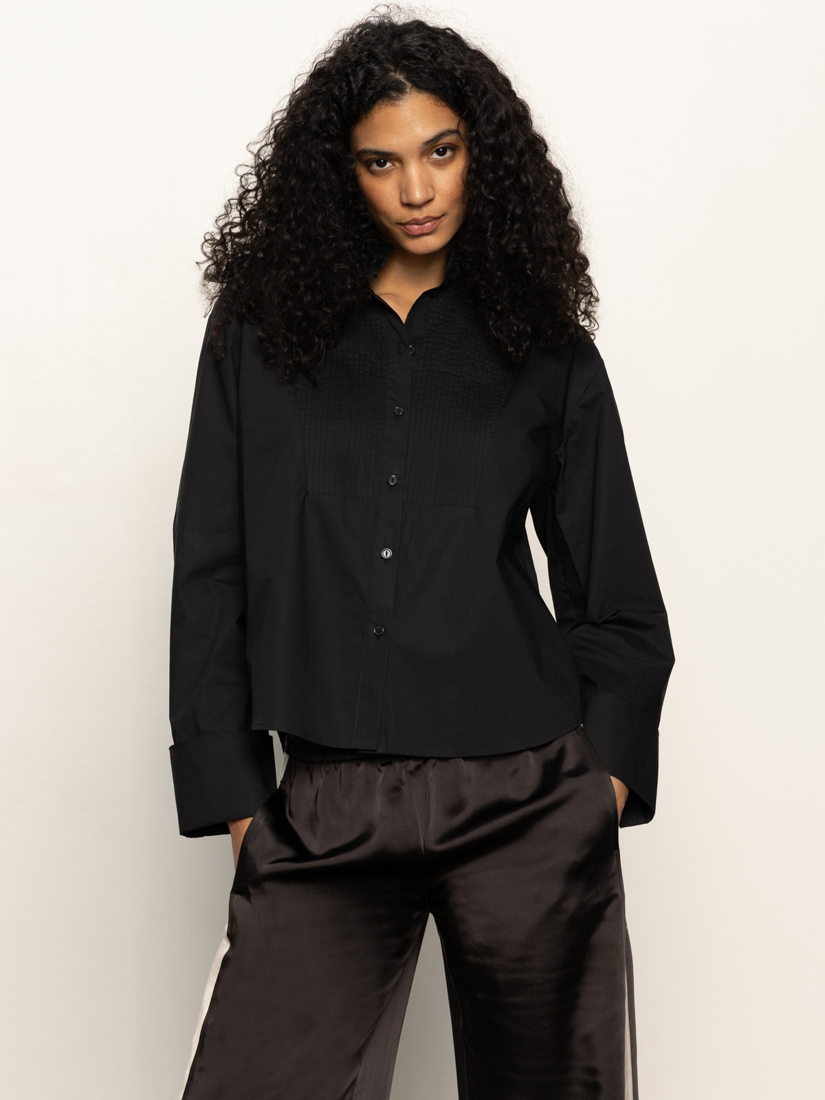 A woman with long curly hair wears the Sanctuary Clothing new era tuxedo shirt in black, paired with dark satin pants, standing against a plain light background.
