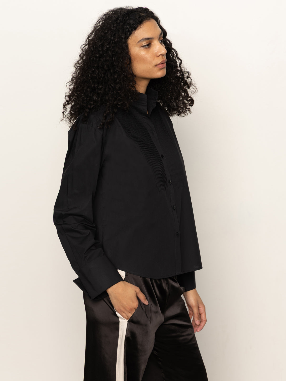 Someone with curly dark hair wears the Sanctuary Clothing new era tuxedo shirt in black, paired with shiny dark pants featuring a white stripe, standing against a plain light background and looking to the side.