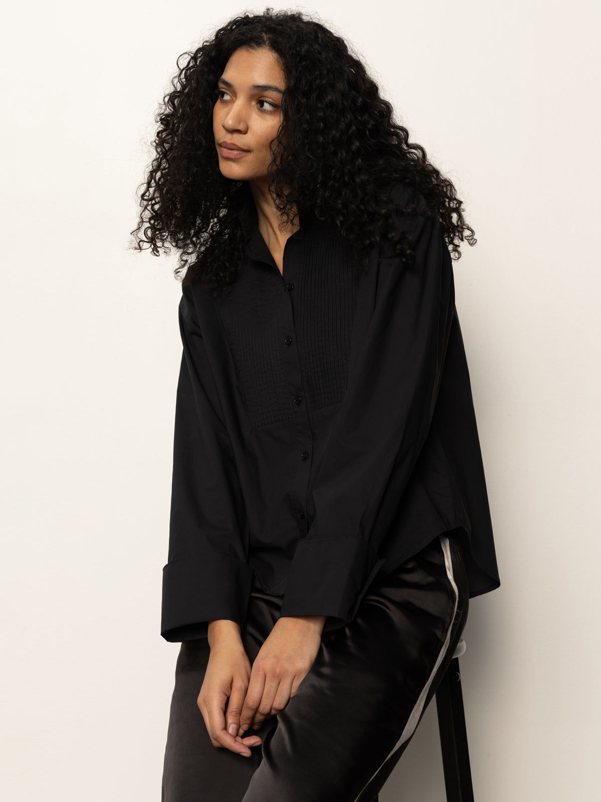 A woman with long, curly hair sits on a stool against a plain background, wearing the Sanctuary Clothing new era tuxedo shirt in black. She looks thoughtfully to the side.