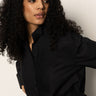 A woman with long, curly dark hair wears the Sanctuary Clothing new era tuxedo shirt in black and looks to her left with a neutral expression, set against a plain light background.