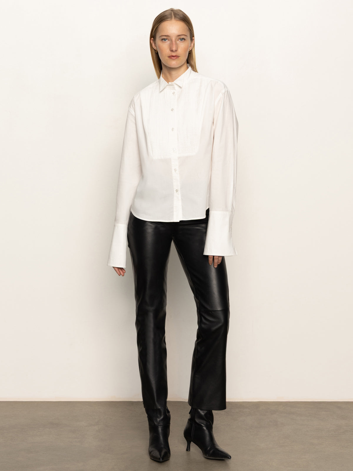 A woman with straight light brown hair and a neutral expression wears the Sanctuary Clothing new era tuxedo shirt in white, paired with black leather pants and black heeled boots, standing against a plain wall.