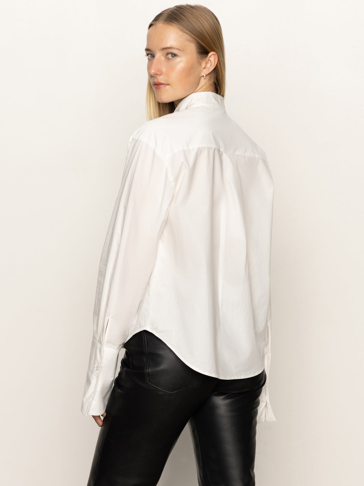 A woman with long blonde hair models the Sanctuary Clothing new era tuxedo shirt in white, paired with black leather pants, standing sideways and looking over her shoulder against a plain light background.