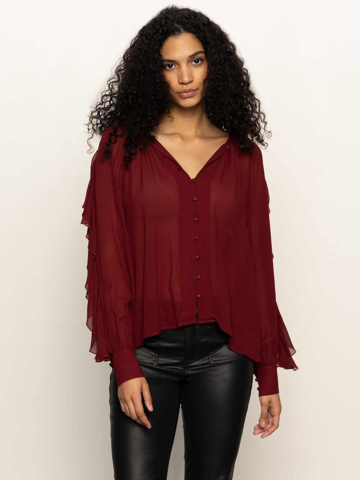 Woman with long curly hair wears a loose, sheer burgundy blouse with buttons and ruffled sleeves, paired with black leather pants, standing against a plain light background.