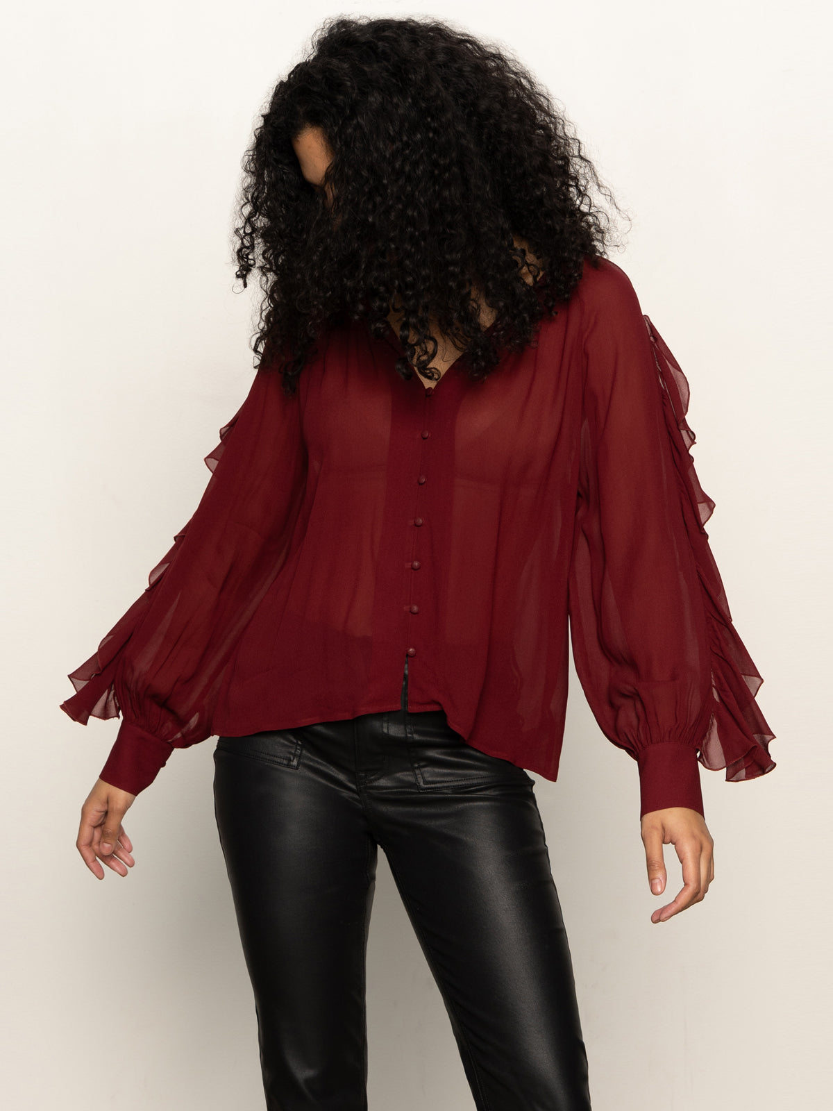 A woman with curly dark hair wears a sheer, long-sleeved burgundy blouse with ruffle details and black leather pants, standing against a plain light background, looking down with her hair covering part of her face.