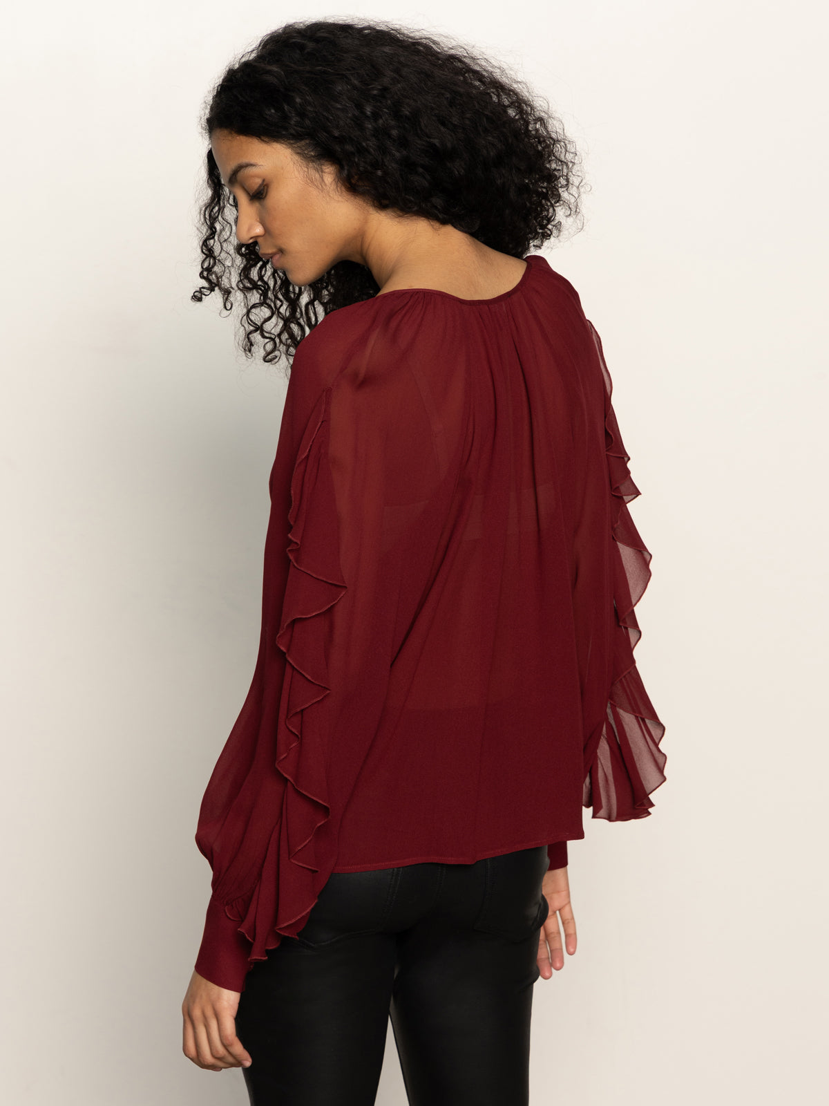 A woman with curly hair wears a sheer, long-sleeve burgundy blouse with ruffles on the sleeves and black pants, standing against a plain light background and looking down over her shoulder.