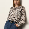 A woman with long, straight blonde hair wears Sanctuary Clothing's "wild dreamer blouse mother leo," a loose animal print top, tucked into high-waisted blue jeans with a black belt, standing against a plain light background and looking at the camera.
