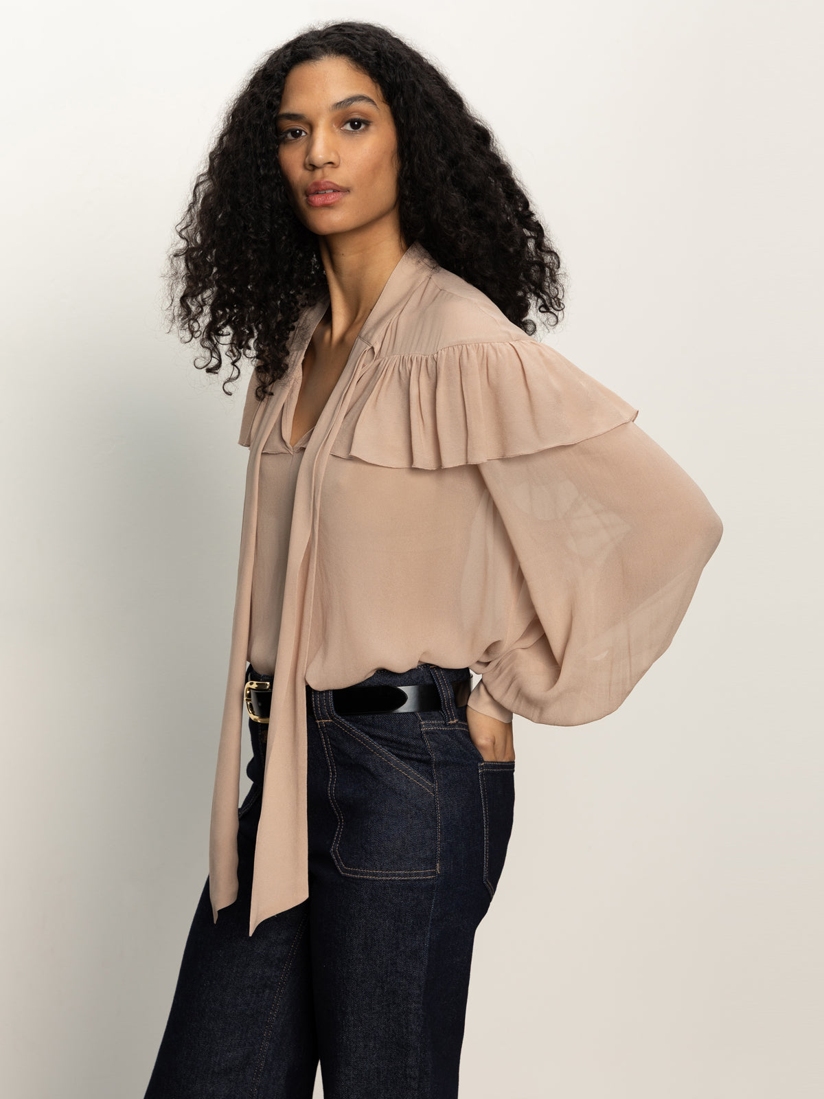A woman with long curly hair wears the Sanctuary Clothing "fade into you ruffle blouse" in fawn, featuring sheer ruffles and loose sleeves, paired with dark blue jeans, posing confidently with one hand in her pocket against a plain backdrop.