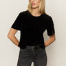 A woman with long blonde hair wears the Sanctuary Clothing "we belong together" velvet top in black, paired with dark gray jeans and a black belt, standing against a plain light background with her hands in her pockets.
