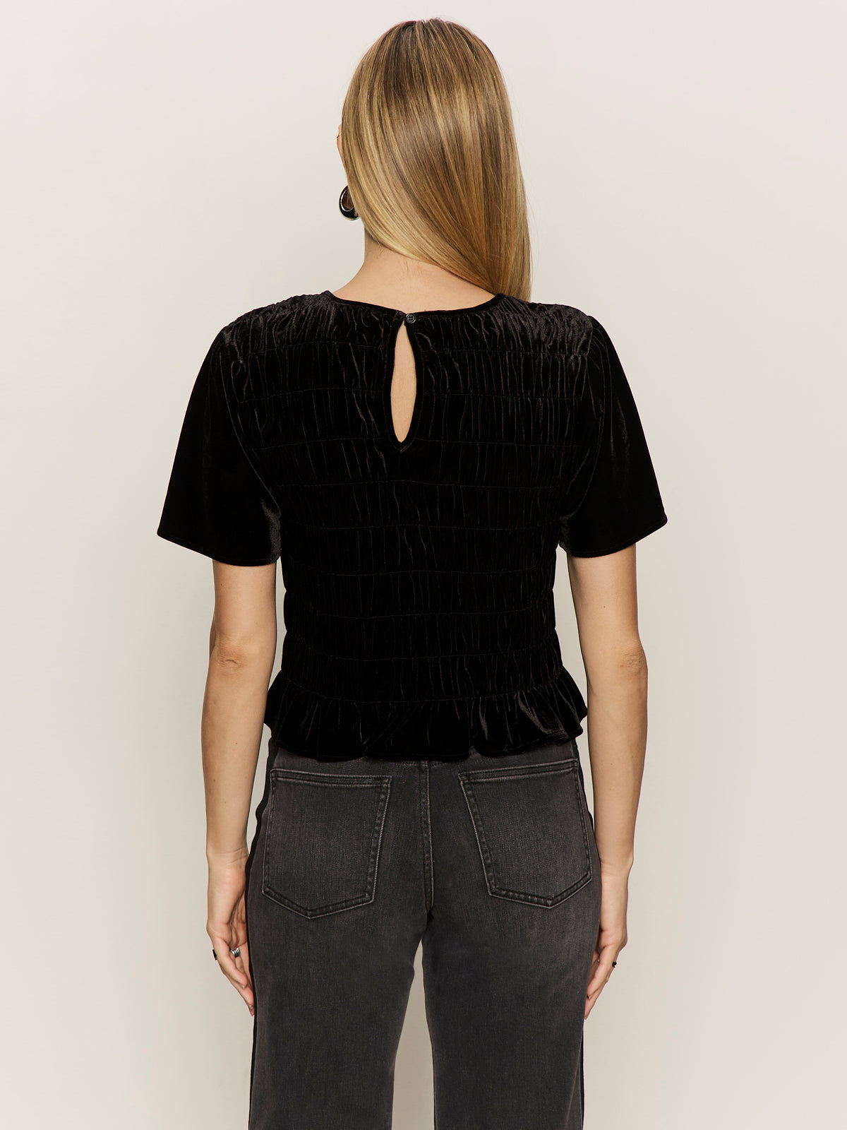 A woman with straight, light brown hair stands with her back to the camera, wearing the Sanctuary Clothing "we belong together" velvet top in black and dark gray jeans against a plain light background.