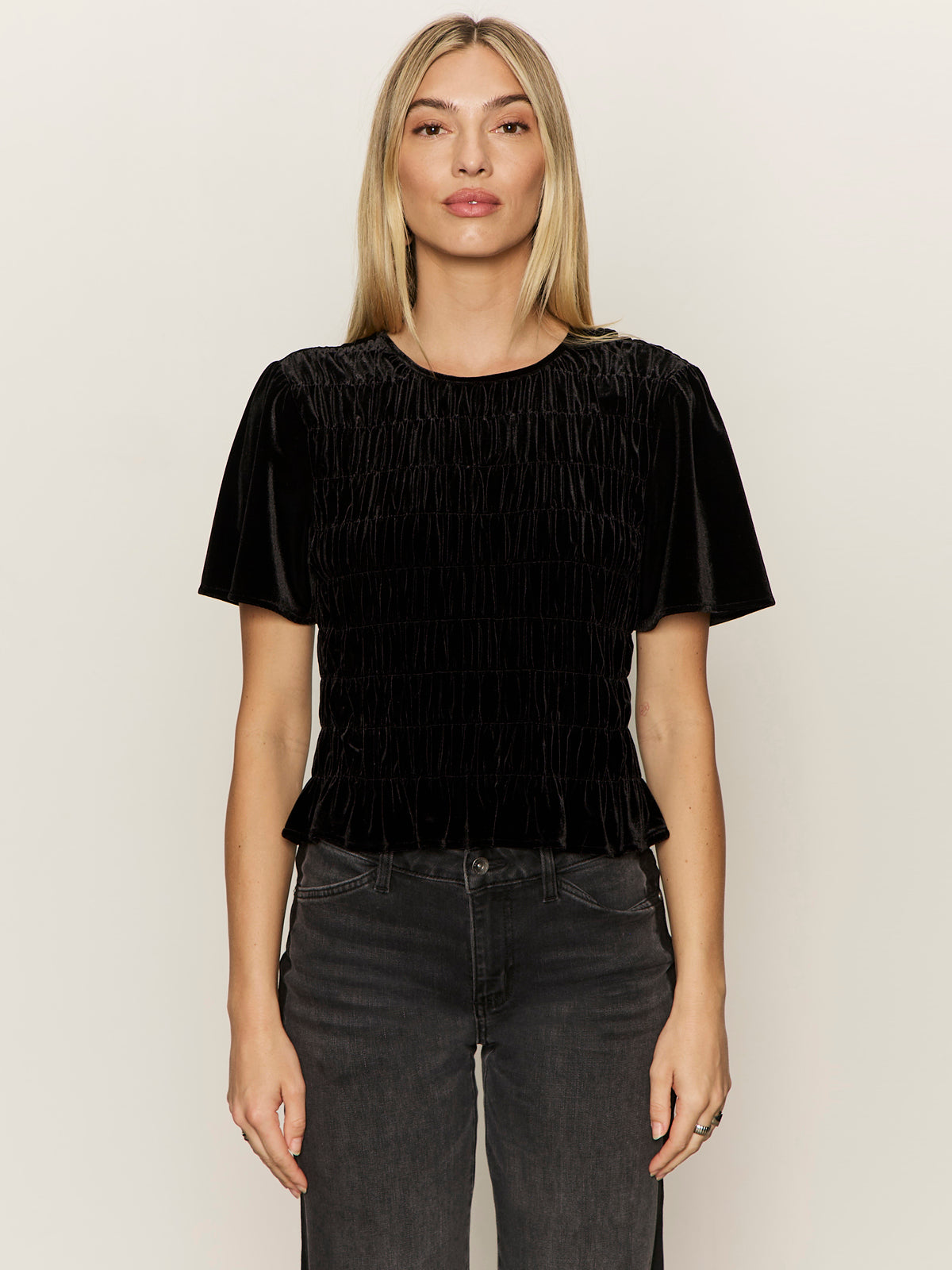 A woman with straight blonde hair faces forward, wearing the black "we belong together velvet top" by Sanctuary Clothing and dark jeans against a plain light background.