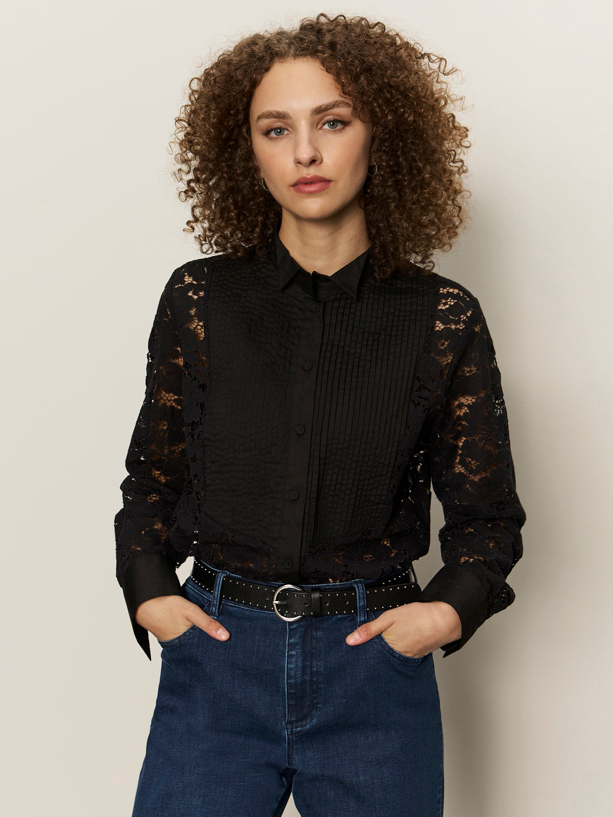 A woman with curly brown hair stands with her hands in her pockets against a light background, wearing the Sanctuary Clothing serendipity lace tuxedo shirt in black and blue jeans.