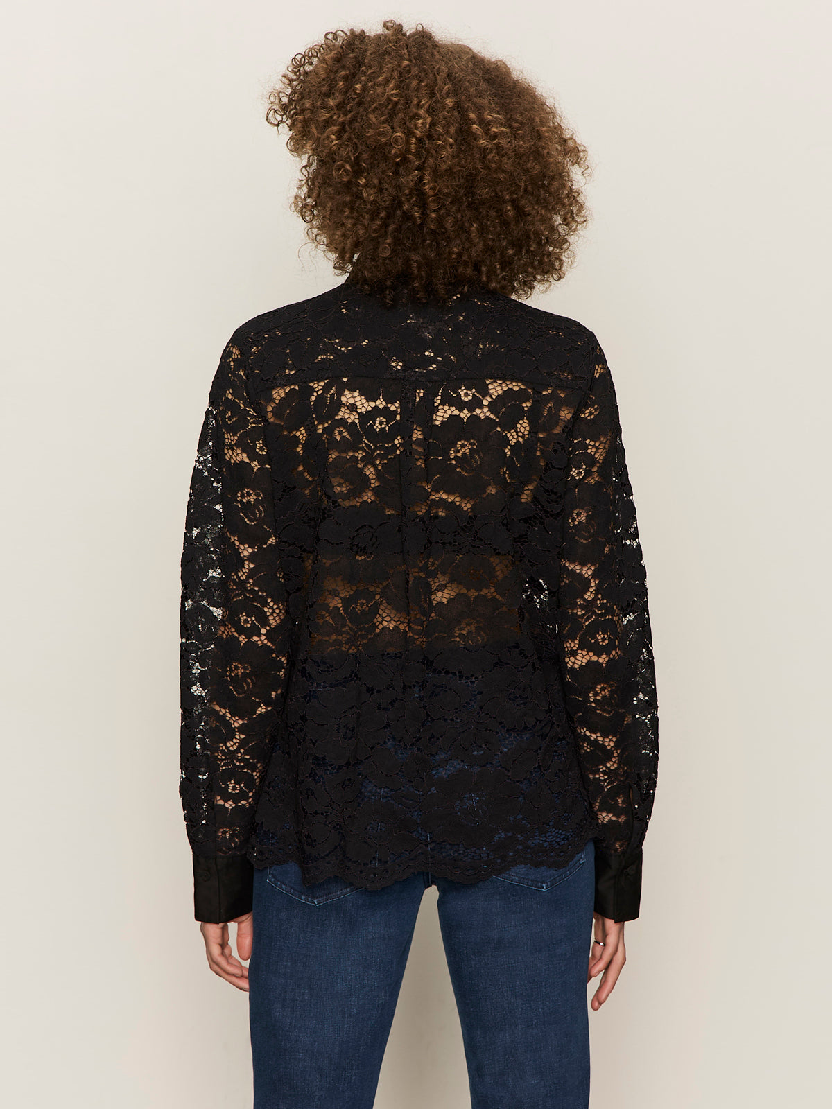 A person with curly hair is shown from the back, wearing Sanctuary Clothing's serendipity lace tuxedo shirt in black over blue jeans. The long-sleeved, see-through blouse features a delicate floral lace pattern.
