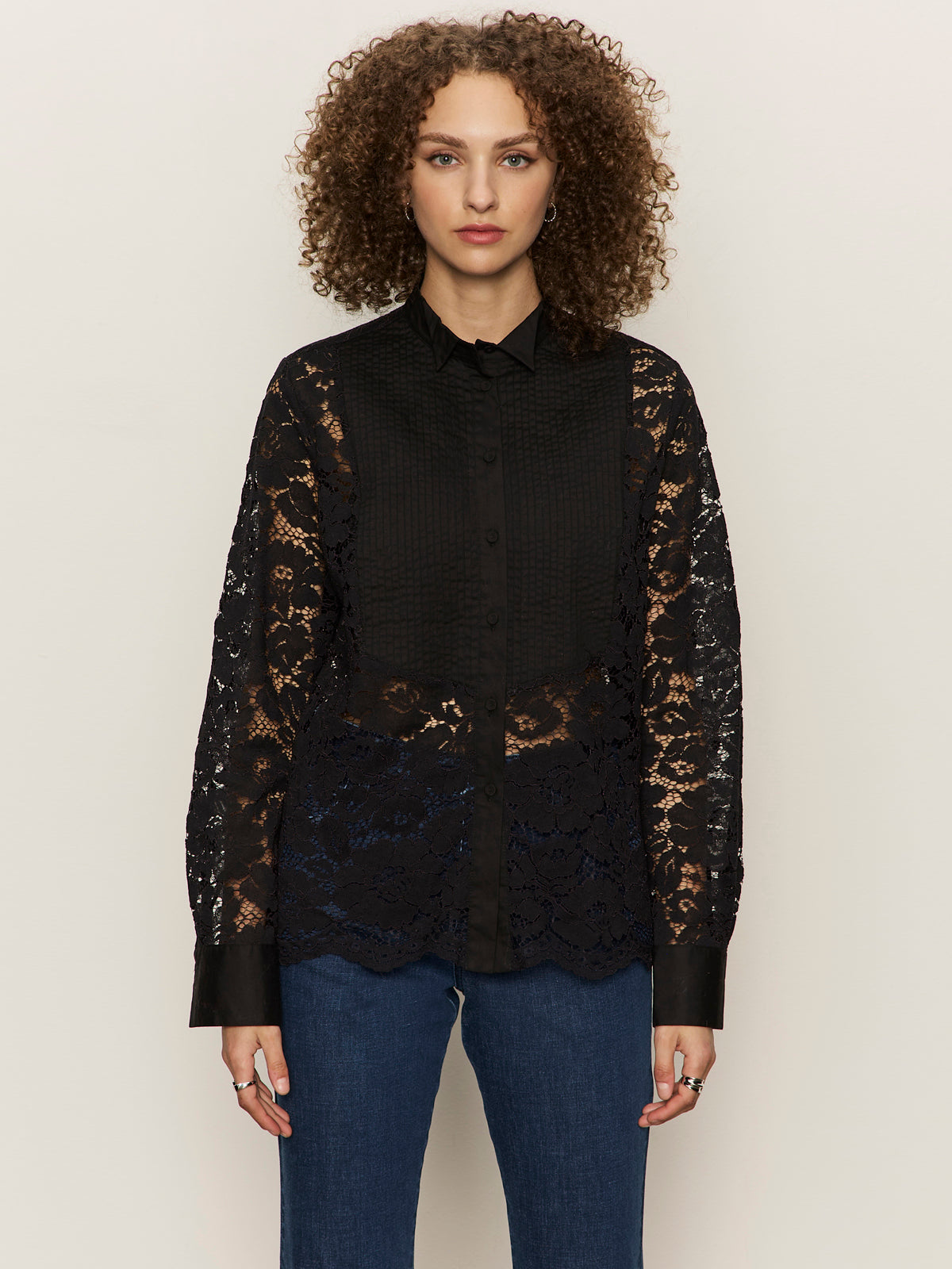 A person with curly hair wears the Sanctuary Clothing serendipity lace tuxedo shirt in black over a black top and blue jeans, standing against a plain beige background.