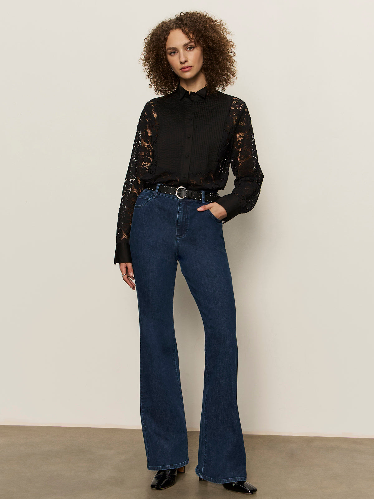 A woman with curly hair wears the Sanctuary Clothing serendipity lace tuxedo shirt in black, paired with blue high-waisted flared jeans, a black belt, and boots. She stands against a light wall with one hand in her pocket.