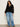 A woman with long curly hair wears the Sanctuary Clothing raye embroidered blouse black extended sizing with long sleeves and blue wide-leg jeans, standing facing forward against a plain light background, one hand relaxed by her side.