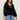 A woman with long curly hair wears the Sanctuary Clothing raye embroidered blouse black extended sizing with floral details and light blue jeans, standing against a plain light background.