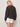 A woman with blonde hair stands facing away in Sanctuary Clothing's joyride relaxed volume blouse rhythmic, a long-sleeved black patterned top, paired with white shorts against a plain light background.