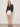 A woman with blonde hair and a neutral expression stands against a plain wall wearing the Sanctuary Clothing joyride relaxed volume blouse rhythmic, paired with white belted shorts and black studded sandals. Her hands rest calmly at her sides.