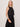 A woman with blonde hair wears Sanctuary Clothing’s “the desired summer crop shell” in black, paired with a matching high-waisted skirt. She accessorizes with gold earrings, a simple necklace, and amber bangles, standing against a plain light background.