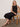 A woman with blonde hair sits on a woven chair, wearing Sanctuary Clothing’s the desired summer crop shell in black. She rests one arm on the chair’s back and looks at the camera with a neutral expression.