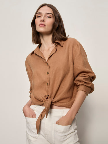 A woman with shoulder-length brown hair wears the Sanctuary Clothing more love linen tie top in toasted coconut, styled with white pants. She stands confidently with hands in pockets against a plain light background, looking at the camera.
