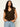 A woman with long, wavy dark hair wears the Sanctuary Clothing outsider embellished shell top in dark cumin black stripe, featuring gold vertical stripes and shoulder details. She stands against a plain light background with one hand in her jeans pocket.