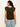 A woman with wavy brown hair wears Sanctuary Clothing's Outsider Embellished Shell Top in dark cumin black stripe, paired with light blue jeans. She stands with her back to the camera against a plain light wall.