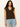 A woman with long wavy dark hair wears the Sanctuary Clothing "outsider embellished shell top" in dark cumin black stripe, styled with light blue jeans, standing against a plain light background.