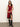 A woman stands against a white wall wearing the Sanctuary Clothing revenge midi satin dress in crimson, featuring a sleeveless design, black lace trim, and thigh-high slit, paired with knee-high black heeled boots.